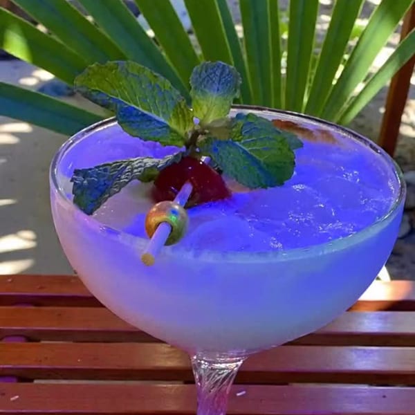 A vibrant blue cocktail, garnished with mint and a cherry, sits on a wooden table.