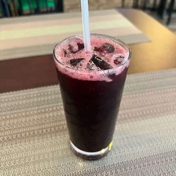 A tall glass of dark red Jamaican sorrel drink with ice and a white straw on a textured placemat.