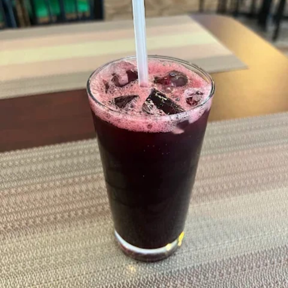 A tall glass of dark red Jamaican sorrel drink with ice and a white straw on a textured placemat.