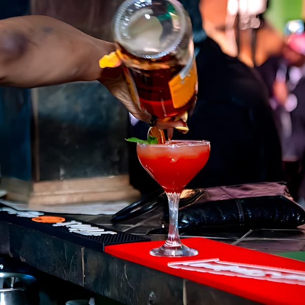 A bartender pours a dark liquid from a bottle into a red cocktail garnished with mint and orange.