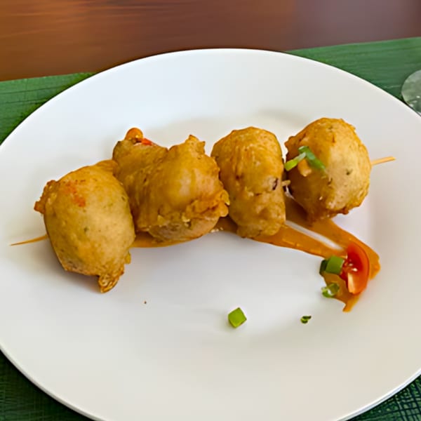Four golden-brown Jamaican fritters on skewers, garnished with sauce, tomato, and green onions.